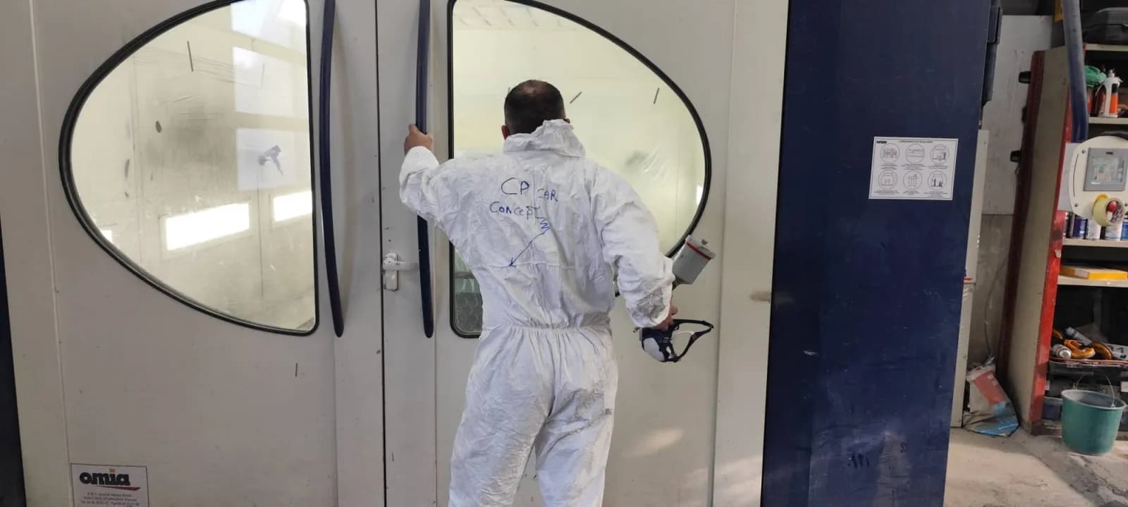 Philippe Cureau de dos en combinaison marquée CP CAR CONCEPT, pistolet de peinture en main, devant la cabine