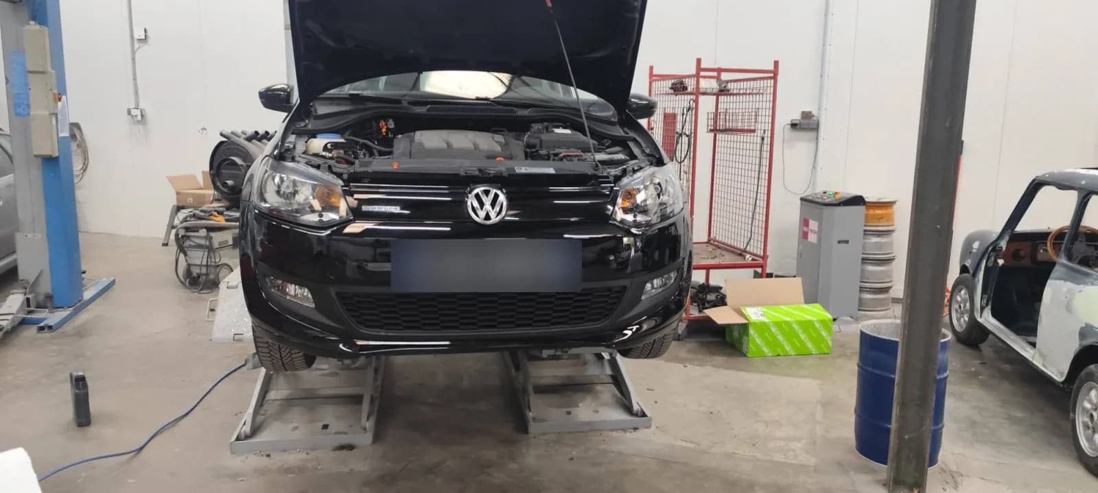 Volkswagen Polo noire de face, avant remonté avec ses deux phares et le pare-chocs, capot ouvert, plaque masquée